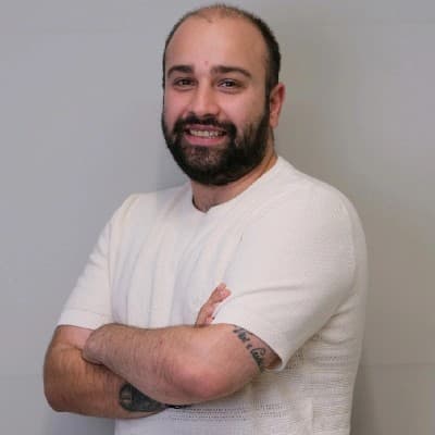 Portrait of İlkan Geldigitti, smiling, arms crossed, in a light knit shirt